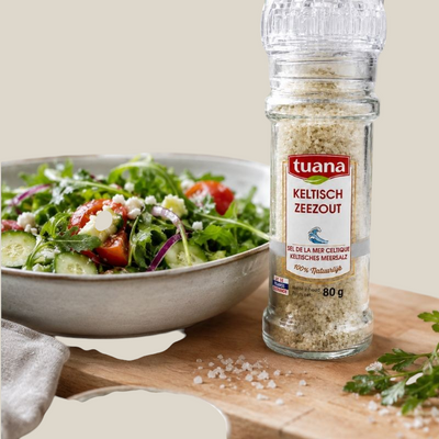 TuanaShop BV Keltisch zeezout in een glazen molen naast een verse salade op een houten snijplank